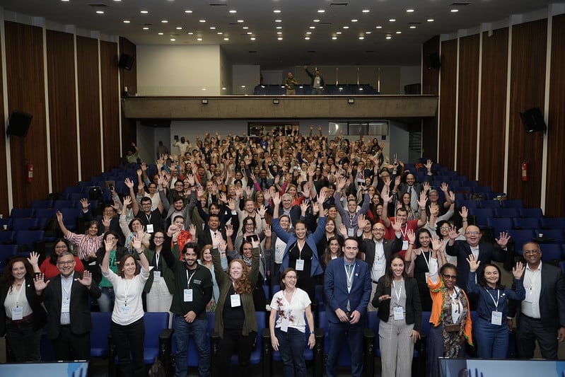 Large group of conference participants gathered in an auditorium in Brasília, smiling and raising their hands during the 1st Americas Regional Meeting on Antimicrobial Resistance.