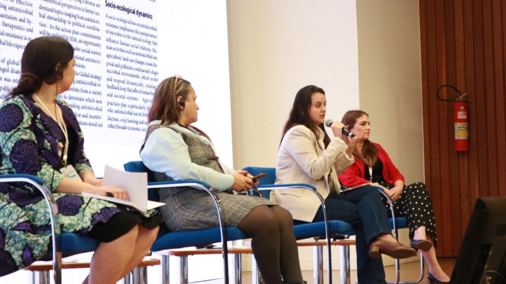 Panel of speakers seated on stage during a discussion on antimicrobial resistance, with one speaker holding a microphone and presentation slides displayed behind them.
