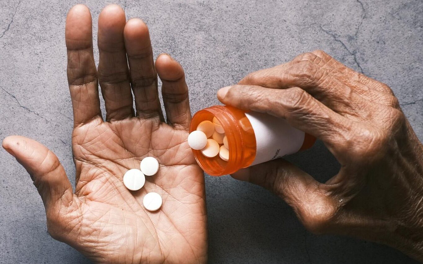 An older adult pours white pills from an orange prescription bottle into their open hand.